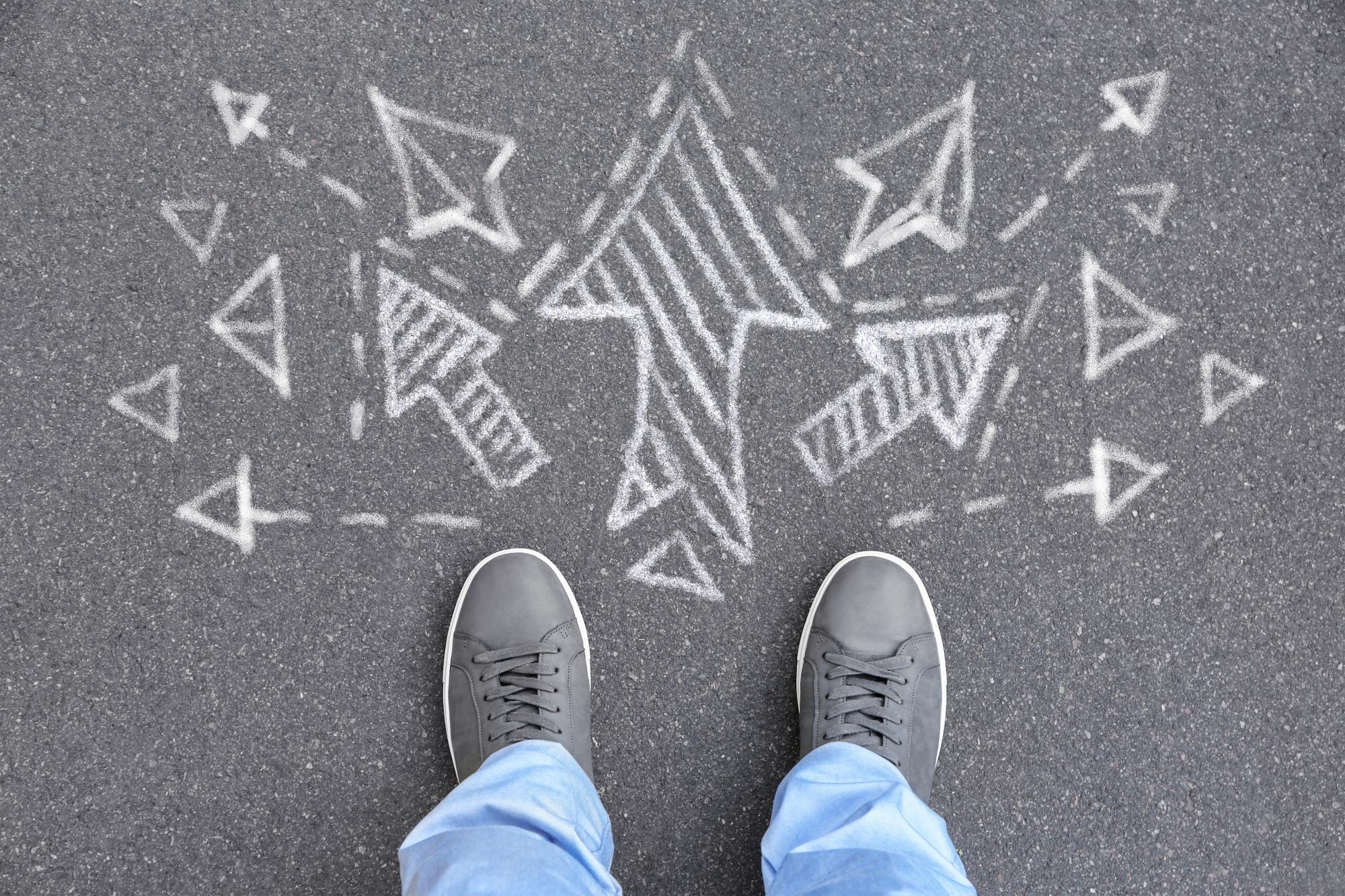 Photo of person's looking down at their shoes in front of a chalk drawing with multiple arrows pointing in different directions. 